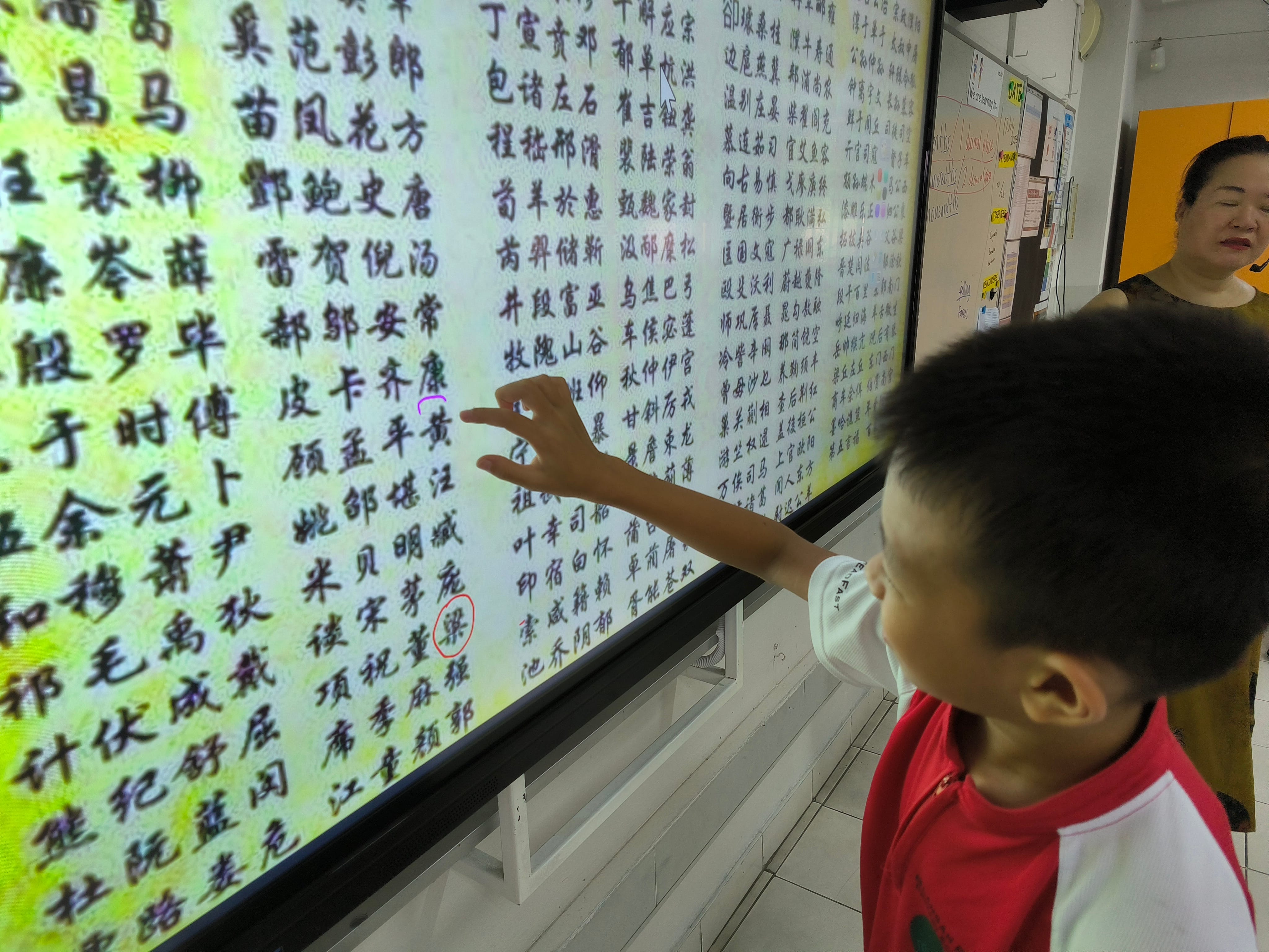 A young boy in a red shirt points at a large screen displaying Chinese characters, with a woman standing nearby.